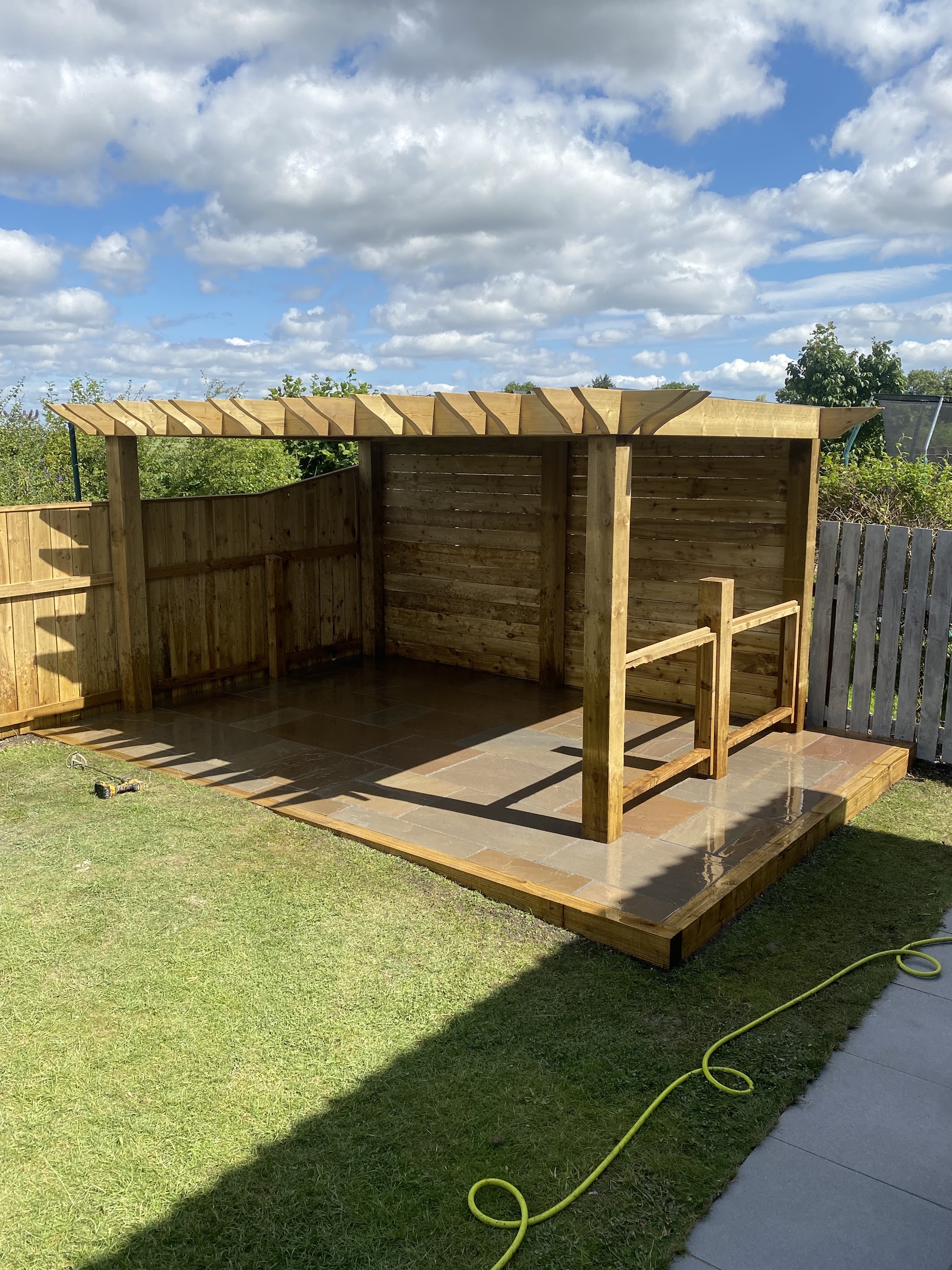 A timber pergola over a decked seating area with a handrail, under a blue sky