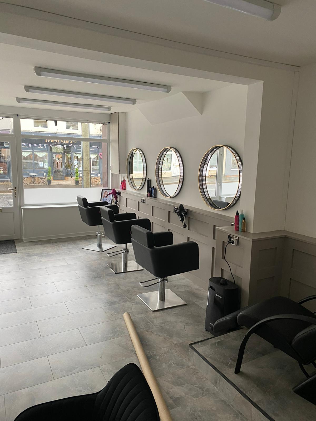 A fitted hair salon with round mirrors above a panelled wall and black styling chairs